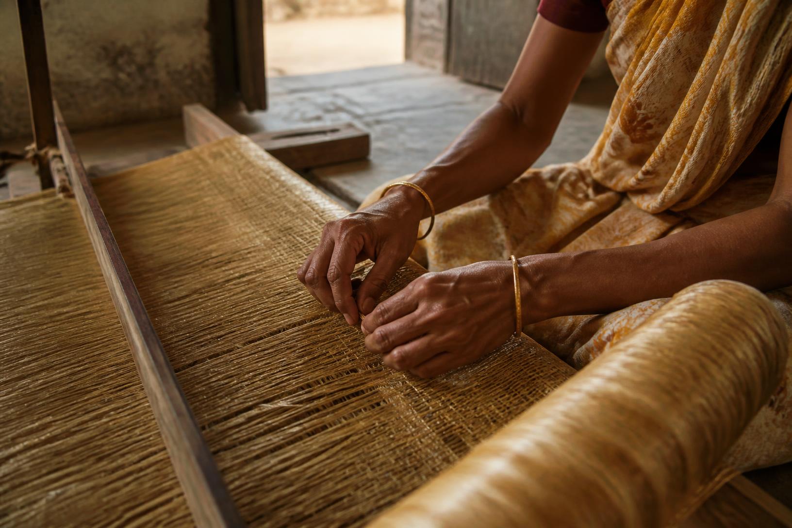 A weaver's hands working golden jute on a floor loom